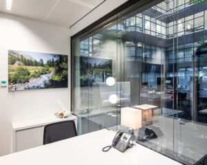 Modern kantoorinterieur met een bureau, telefoon en lamp, uitkijkend op een glazen atrium met zicht op andere kantoorruimtes.
