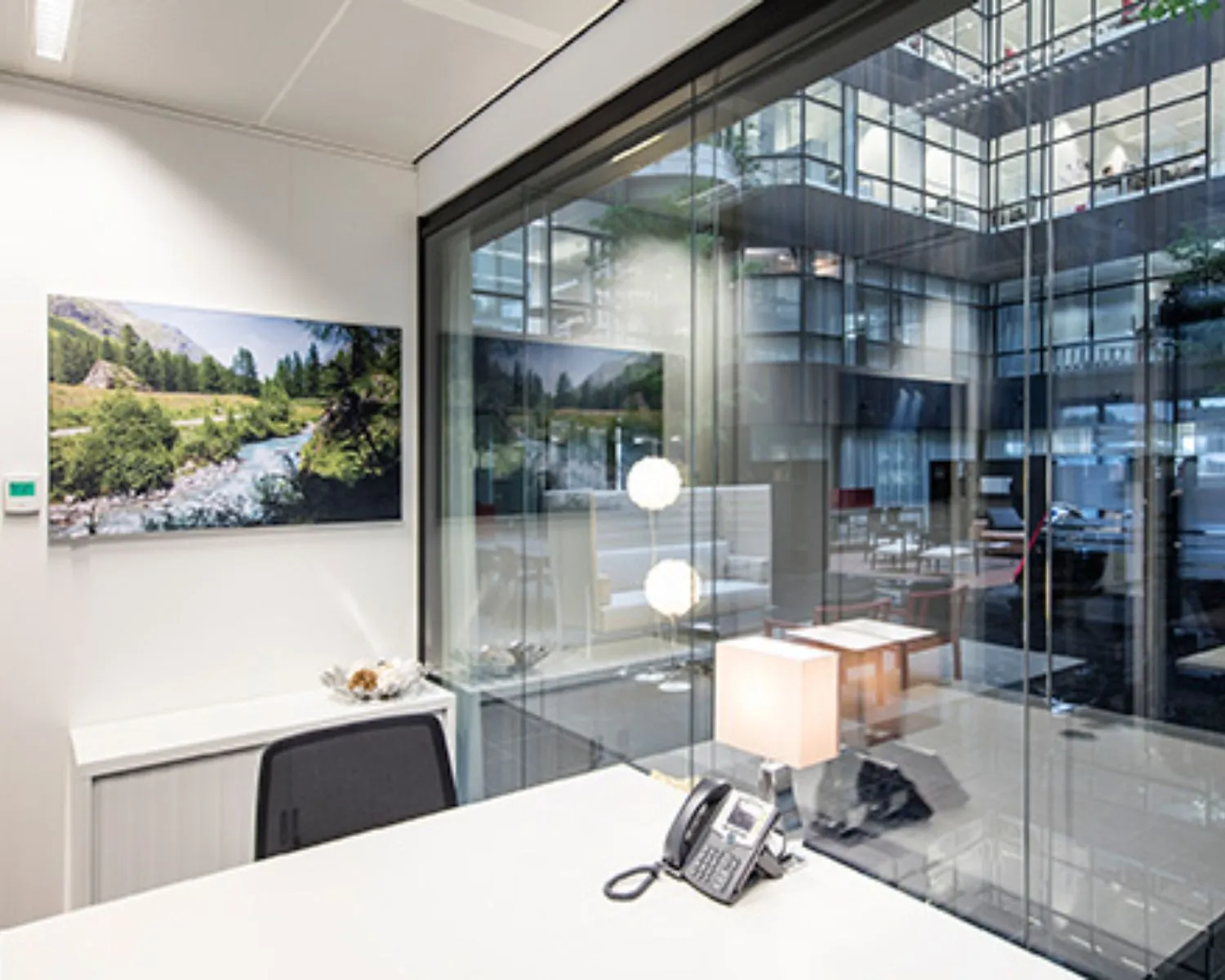 Modern kantoorinterieur met een bureau, telefoon en lamp, uitkijkend op een glazen atrium met zicht op andere kantoorruimtes.