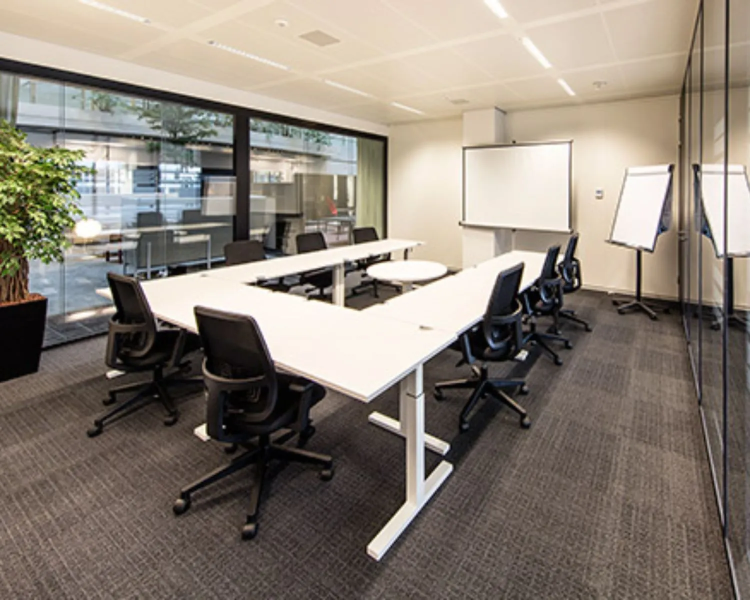 Vergaderruimte met een U-vormige opstelling van bureaus, zwarte bureaustoelen, een whiteboard en flip-overs.