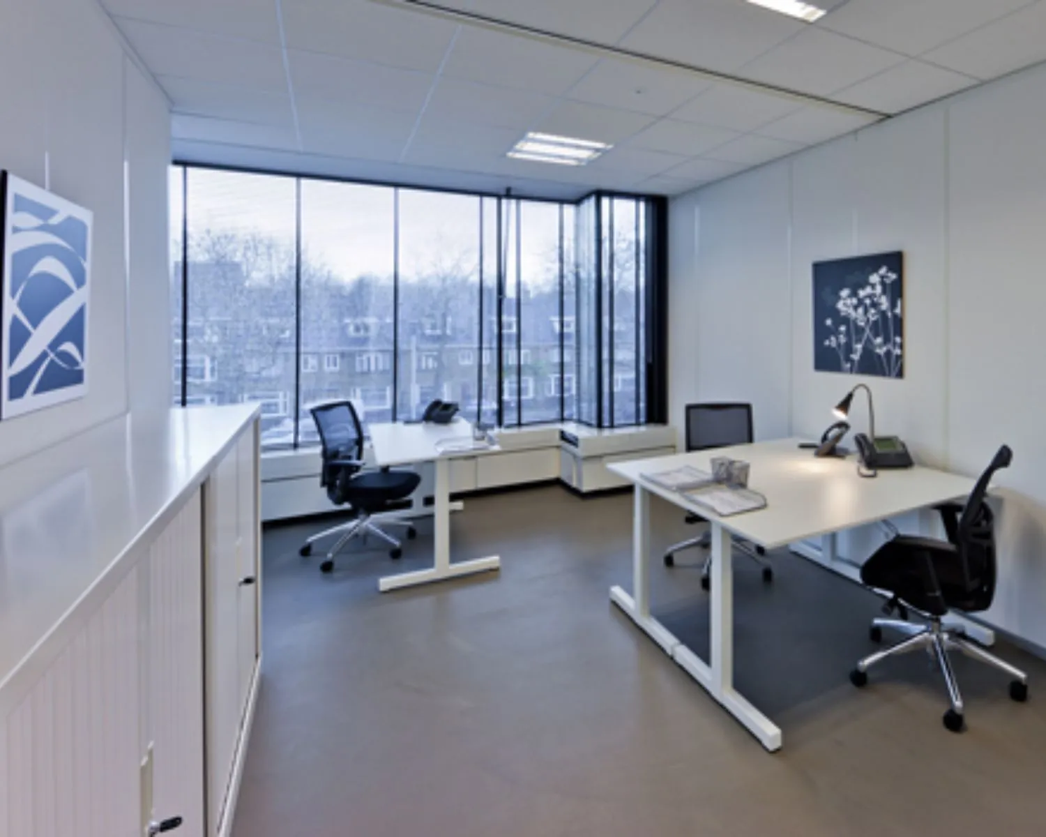 Modern ingericht kantoor met twee bureaus, bureaustoelen en grote ramen met uitzicht op een woonwijk.