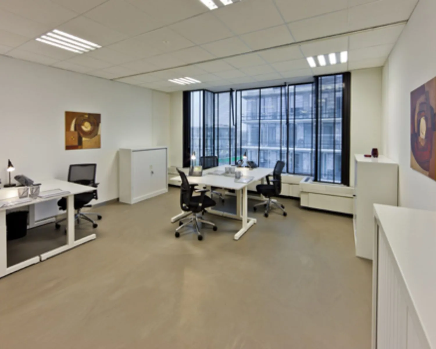 Modern kantoor met vier bureaus, bureaustoelen, grote ramen en abstracte kunst aan de muren.