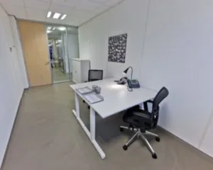 Klein, modern kantoor met een wit bureau, bureaustoel, bureaulamp en telefoon, gelegen in een minimalistische ruimte met glazen deur.