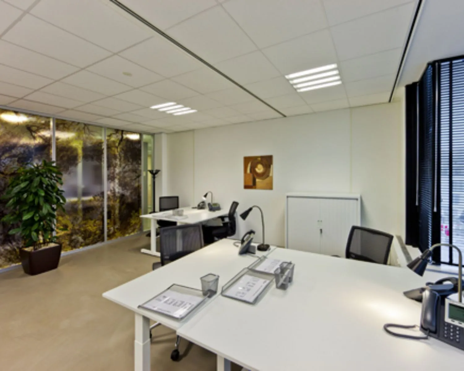 Modern ingericht kantoor met vier bureaus, strakke witte meubels en een natuurprint op de glazen wand.