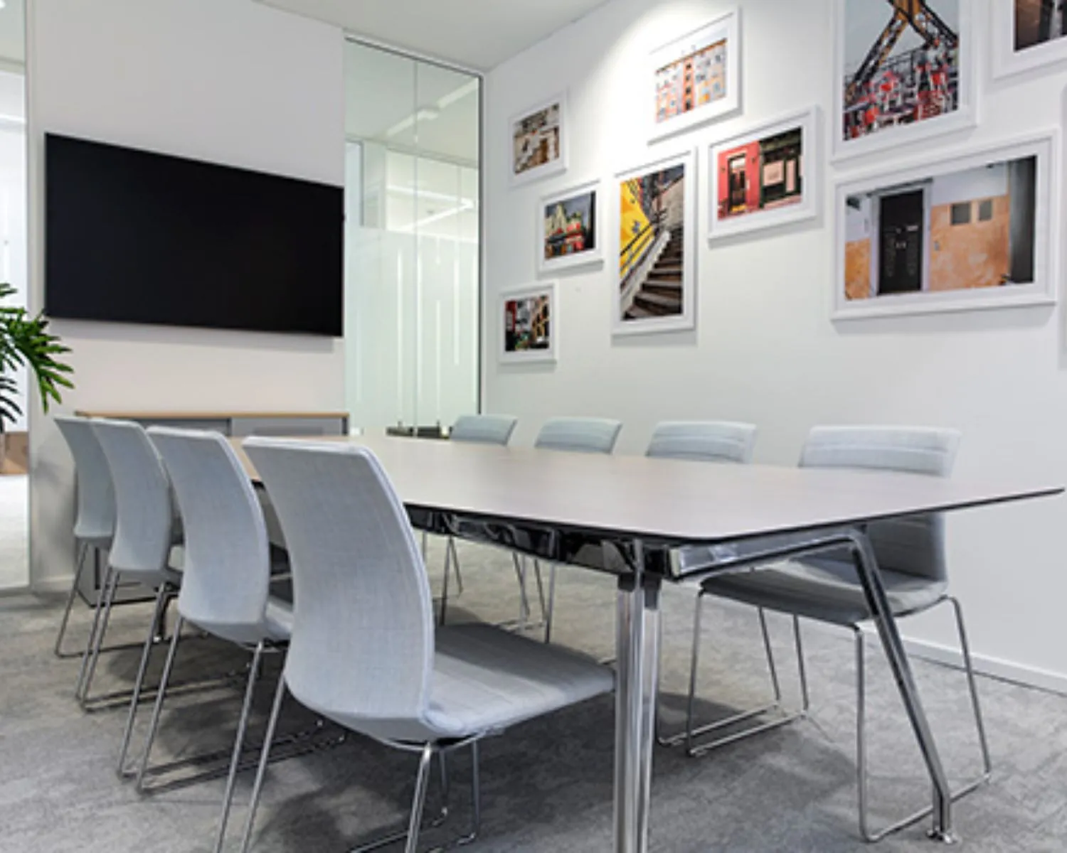 Vergaderruimte met moderne grijze stoelen, een houten tafel, een flatscreen en kleurrijke foto's aan de muur.