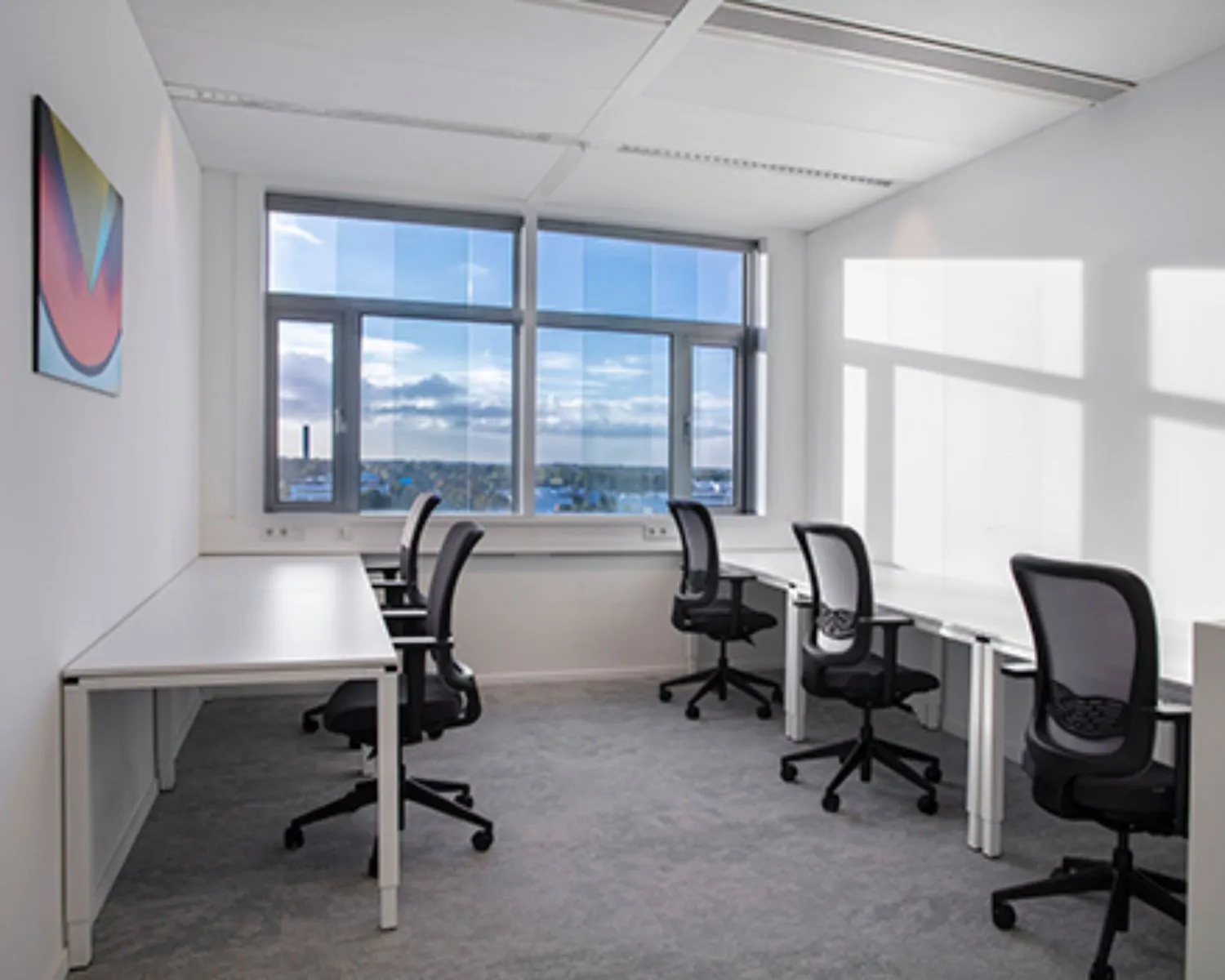 Lichte, moderne kantoorruimte met vijf bureaus en bureaustoelen bij grote ramen.