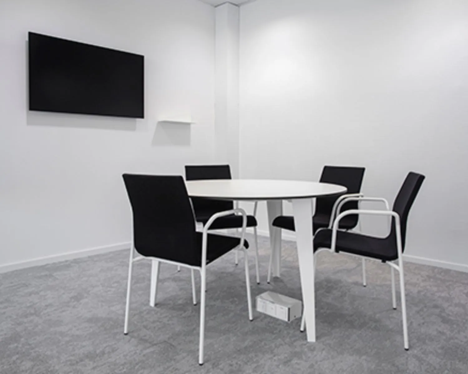Vergaderruimte met een ronde witte tafel, vier zwarte stoelen en een flatscreen aan de muur.