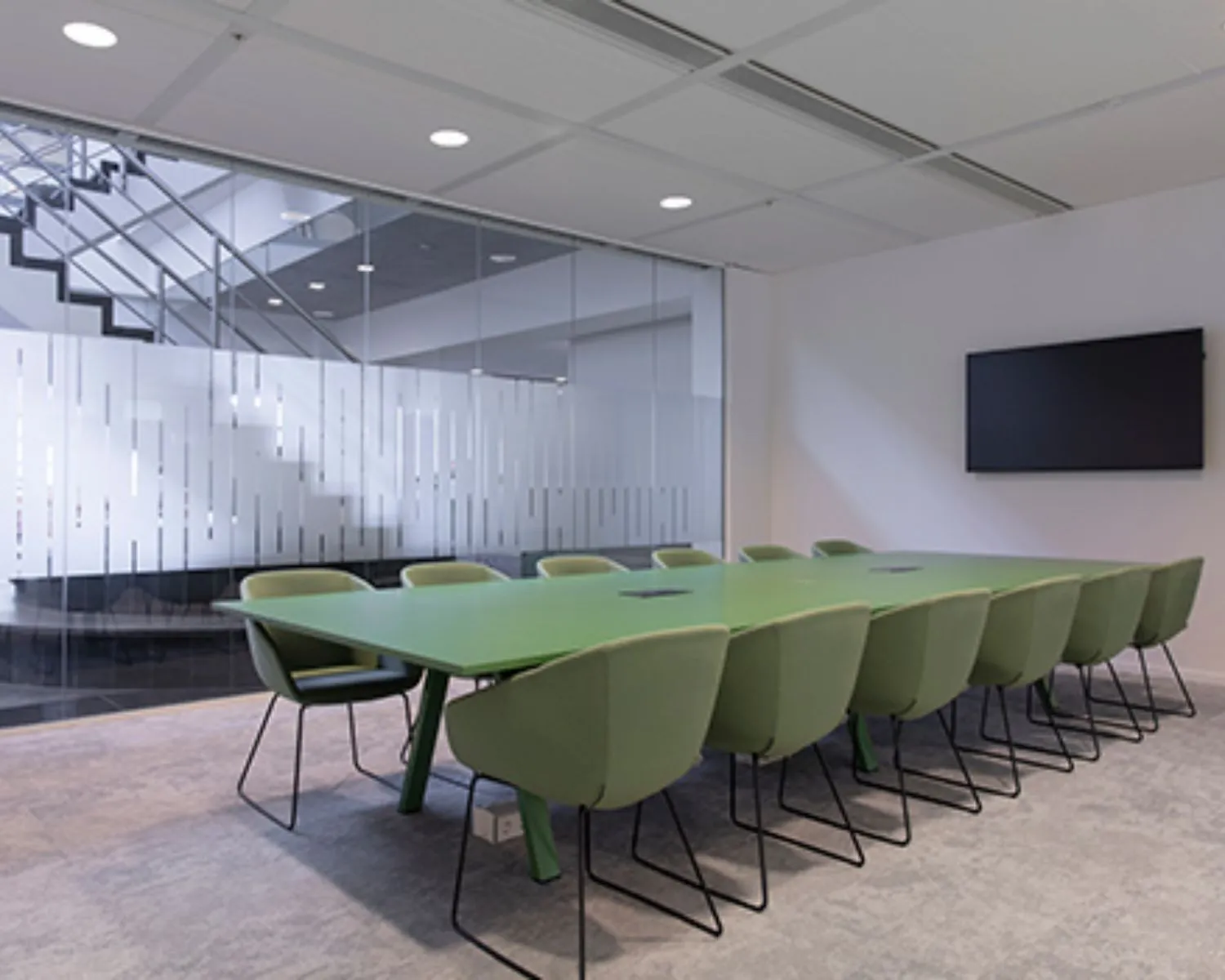 Moderne vergaderruimte met een lange groene tafel, groene stoelen en een flatscreen aan de muur.