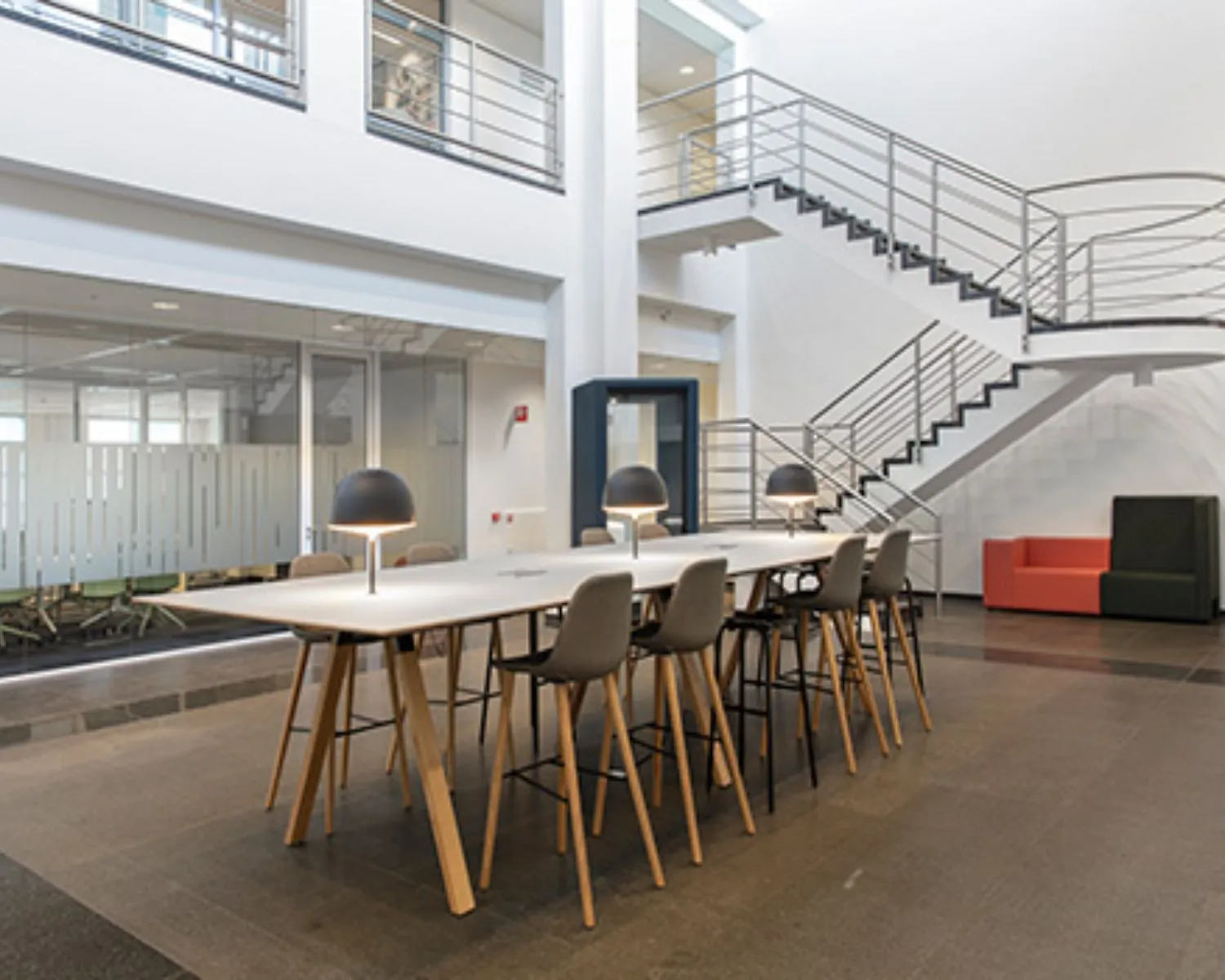 Moderne kantoorlobby met lange vergadertafel, designlampen, zitmeubilair en een trap naar de bovenverdieping.