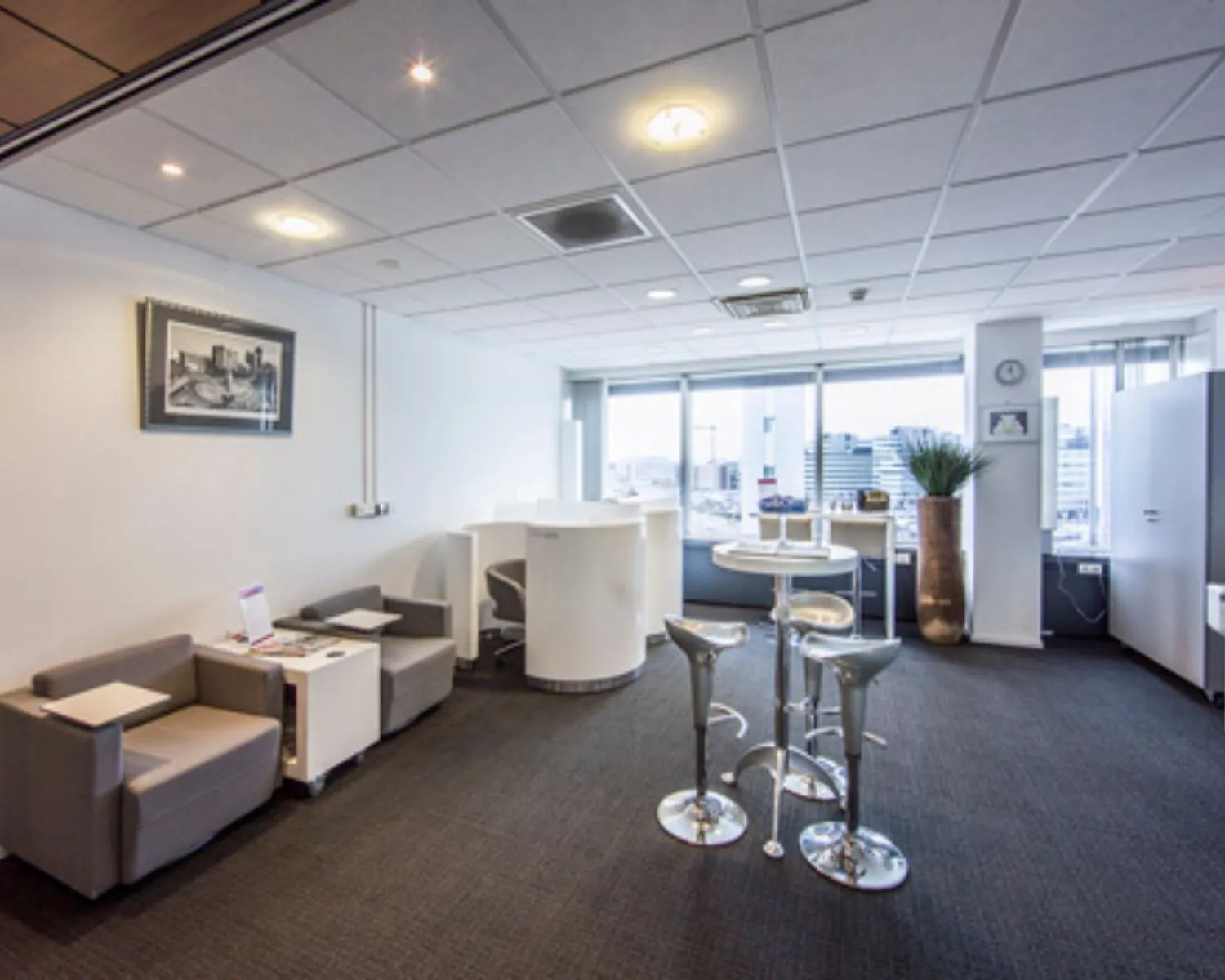 Moderne en lichte kantoorlounge met zitgedeelte, ronde statafel en uitzicht op een stedelijke skyline.