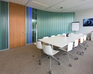 Moderne vergaderruimte met een lange witte tafel, witte stoelen op wieltjes en een flip-over tegen een groene glazen wand.