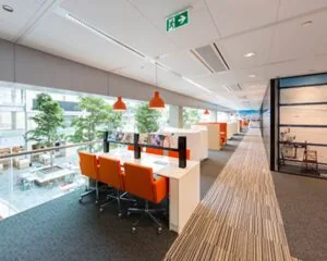 Moderne kantoorruimte met werkplekken, oranje stoelen en hanglampen, grenzend aan een open atrium met veel daglicht.
