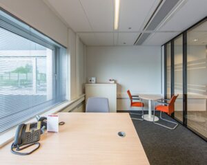 Modern ingericht kantoor met een bureau, vergadertafel en twee oranje stoelen bij een glazen wand.