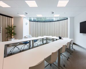 Moderne vergaderruimte met U-vormige opstelling van witte tafels en stoelen, grote ramen en een flatscreen aan de muur.