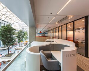 Moderne kantoorruimte met een afgeronde werkplek genaamd "thinkpod" en uitzicht op een open atrium met bomen en zitplaatsen.