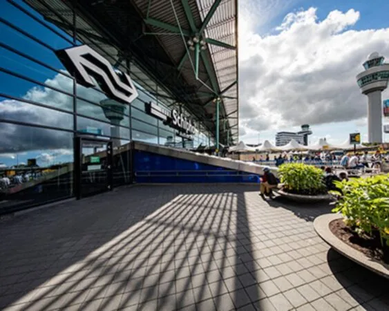 De ingang van station Schiphol met het NS-logo en de verkeerstoren van Schiphol op de achtergrond.