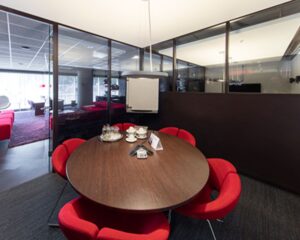 Vergaderruimte met een ronde houten tafel, rode stoelen en een flip-over, grenzend aan een open lounge met glazen wanden.