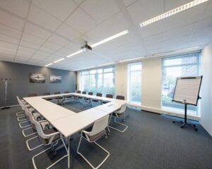 Moderne vergaderruimte met U-vormige opstelling, conferentiestoelen, flipover en grote ramen.