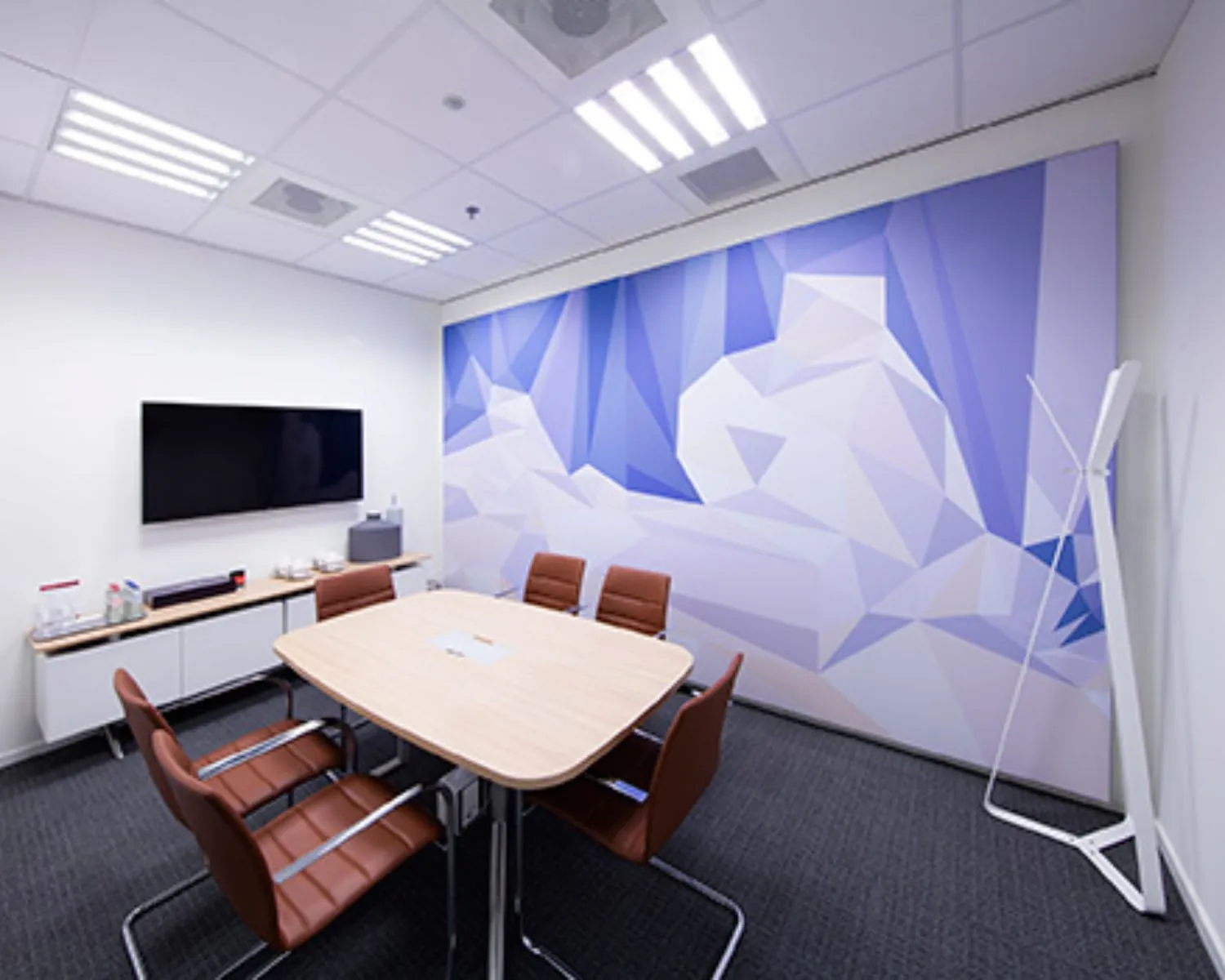 Vergaderruimte met zes bruine stoelen, een ovale tafel, een muurvullende abstracte muurschildering en een flatscreen tv.