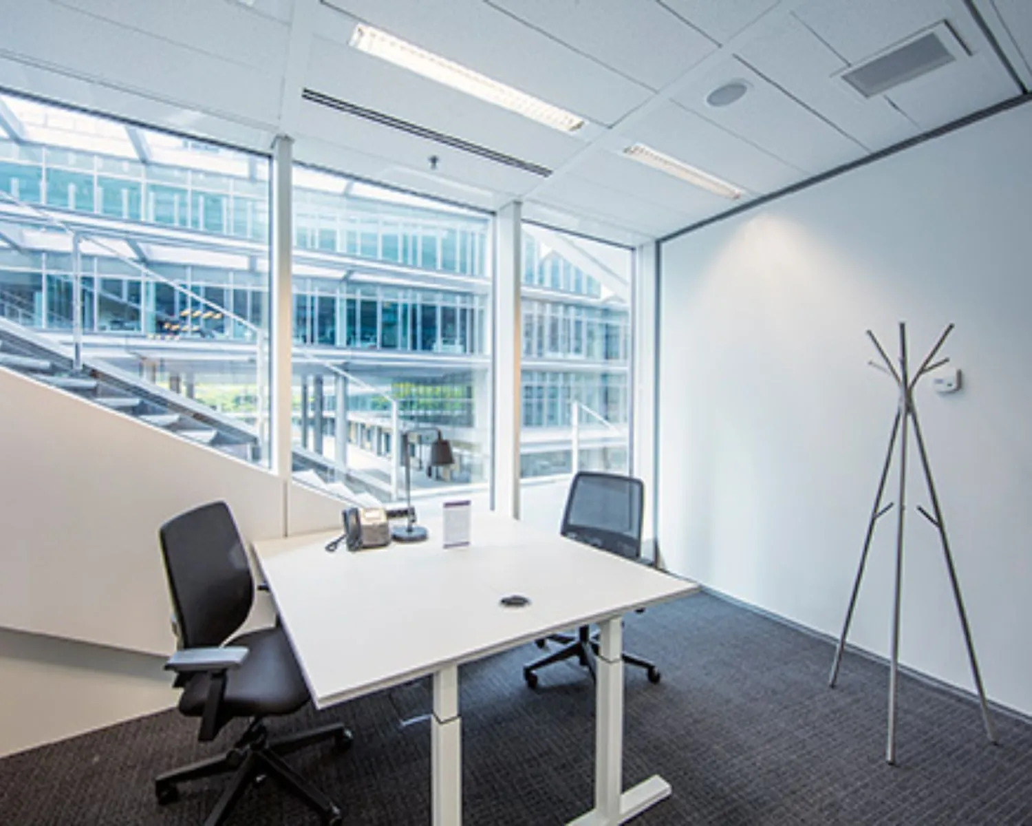 Kleine, moderne kantoorkamer met twee bureaustoelen, een telefoontoestel en groot raam met uitzicht op een glazen kantoorgebouw.