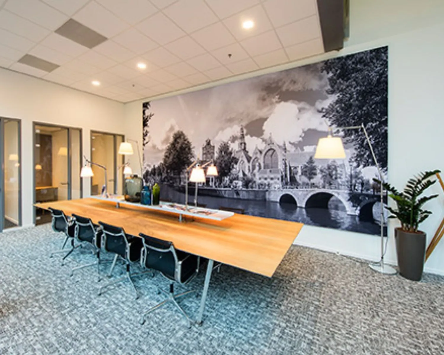 Moderne vergaderruimte met een lange houten tafel, bureaulampen en een grote zwart-wit muurfoto van een stad met brug en historische gebouwen.