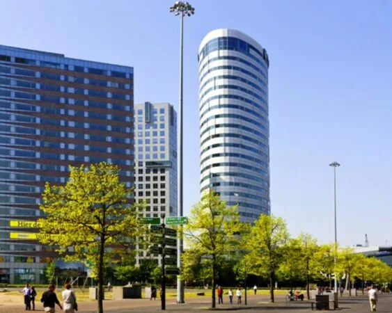 Moderne kantoorgebouwen en wandelaars op een zonnig plein met jonge bomen in een stedelijke omgeving.