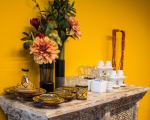 Een houten tafel met kleurrijk Marokkaans aardewerk, bloemen in vazen en koffiekopjes tegen een gele muur.