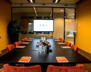 Vergaderruimte met lange tafel, oranje stoelen, notitieboekjes en groot scherm voor presentaties.