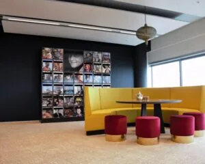 Moderne loungehoek met gele bank, rode krukjes en een wand met portretfoto’s van diverse mensen.