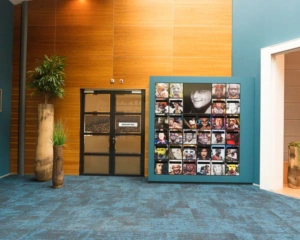 Moderne hal met blauwe vloerbedekking, houten wandpanelen, planten en een kleurrijke fotomuur met portretten naast een deur met het opschrift "DREAMTIME".