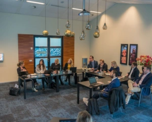 Een groep mensen zit in een vergaderruimte in bespreking rondom een U-vormige tafel met laptops en notitieblokken.