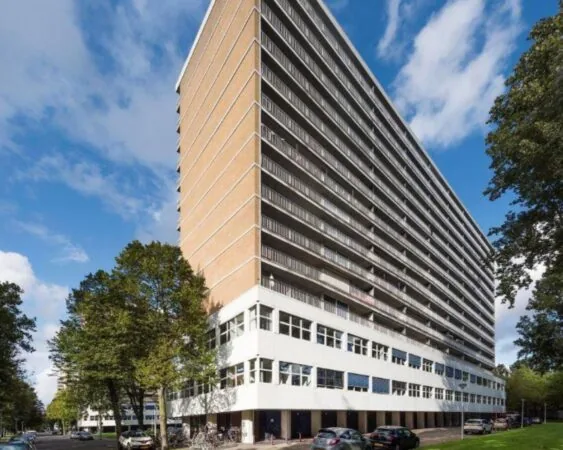Hoog flatgebouw met balkons en een witte begane grond, omringd door bomen en geparkeerde auto's.