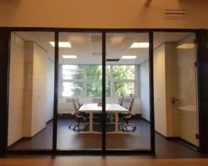 Vergaderruimte met glazen wanden, moderne bureaustoelen en een rechthoekige tafel, met uitzicht op bomen buiten.