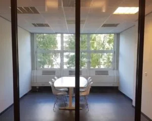 Vergaderruimte met een witte tafel en zes stoelen, gelegen aan een raam met uitzicht op bomen.