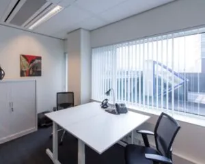 Modern ingericht kantoor met twee bureaustoelen, een wit bureau en een groot raam met verticale jaloezieën.