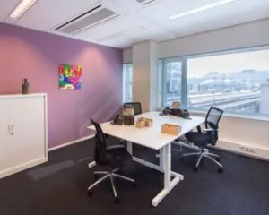 Modern kantoor met twee bureaus, paarse accentmuur, kleurrijk schilderij en uitzicht op een stedelijk treinstation.