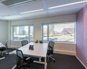 Moderne vergaderruimte met vier bureaustoelen, bureaus en grote ramen met uitzicht op een stedelijke omgeving.