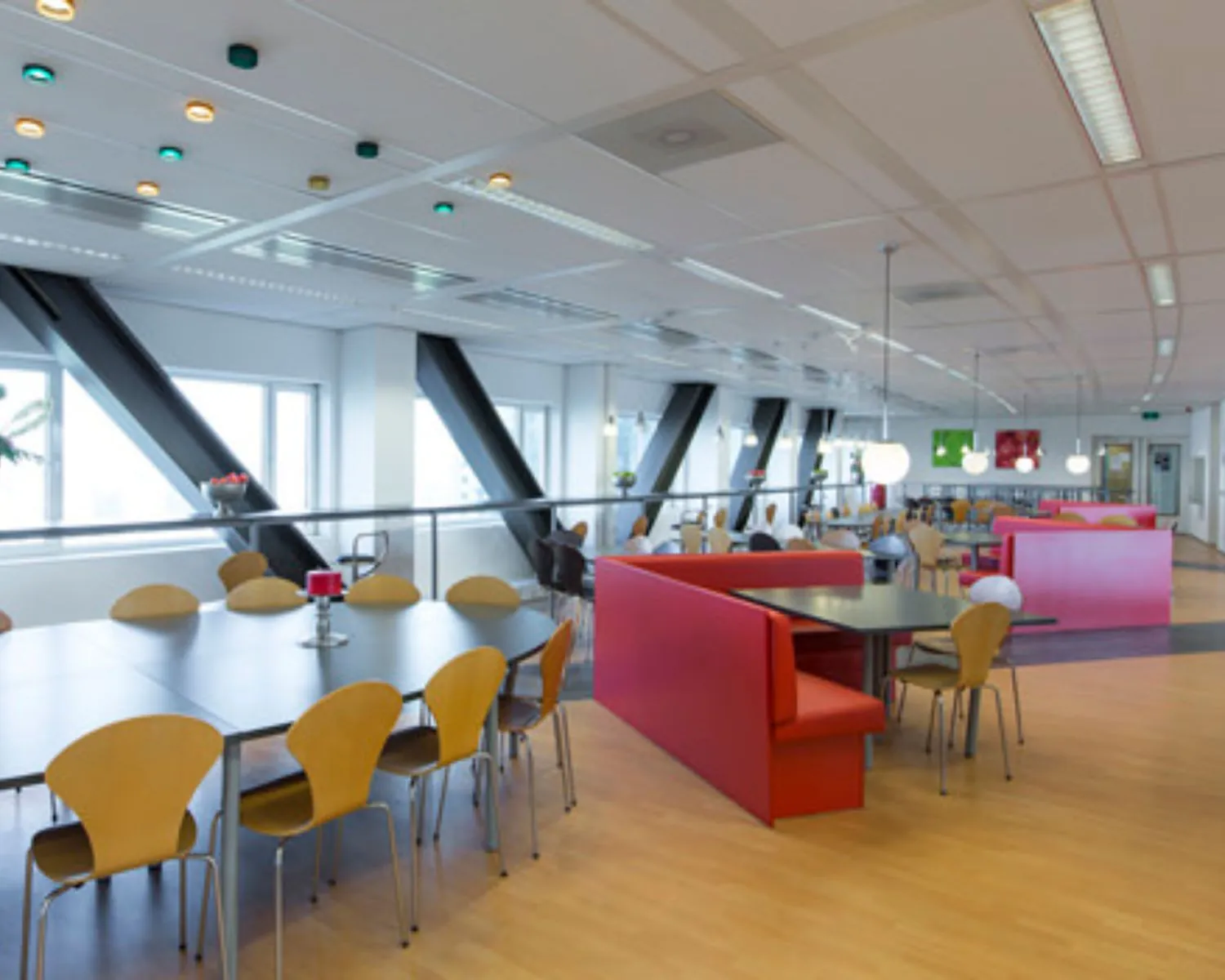 Moderne en kleurrijke bedrijfskantine met houten stoelen, rode zitbanken en grote ramen.