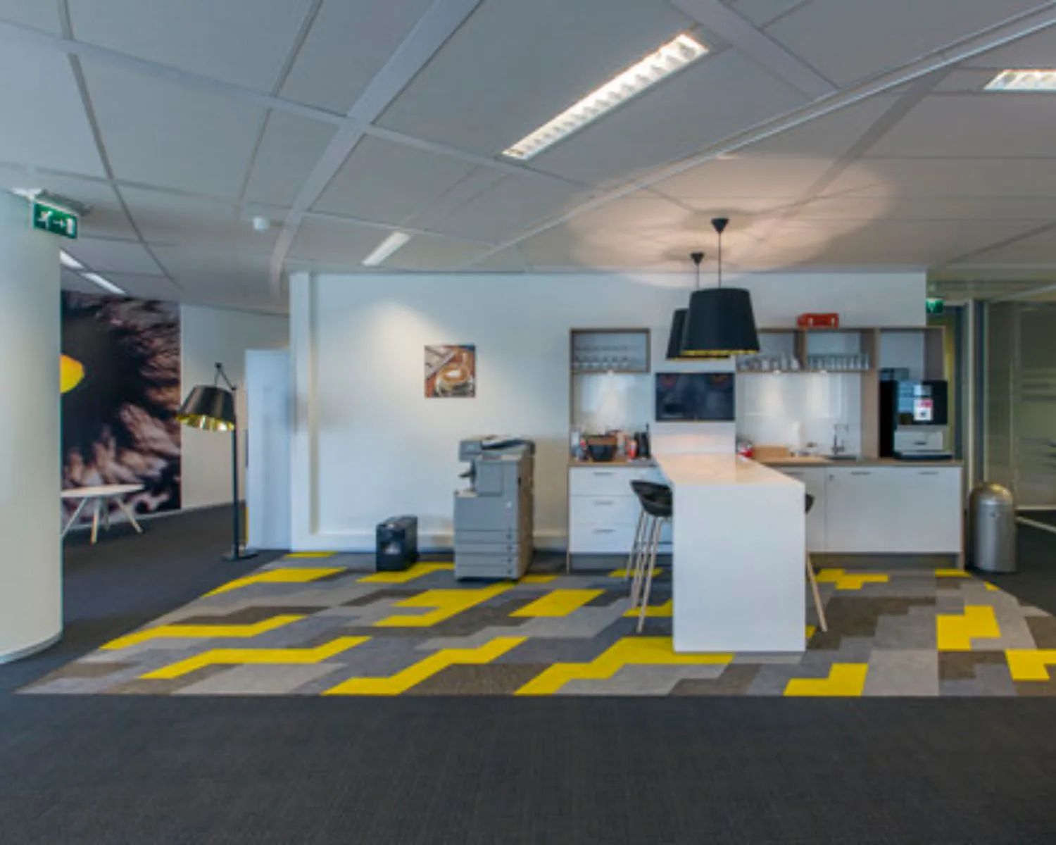 Moderne kantoorruimte met een koffiecorner, printer en opvallend geel-grijs patroon op het tapijt.