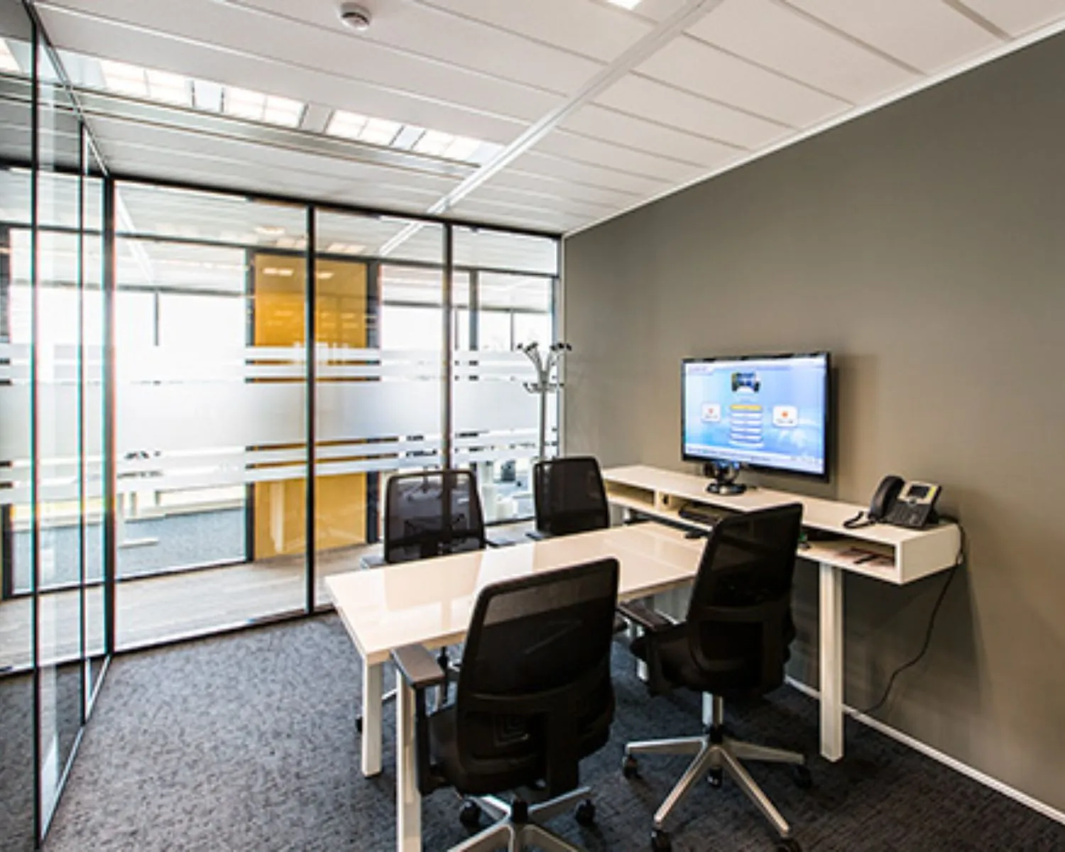 Moderne vergaderruimte met glazen wanden, vier stoelen, een witte tafel en een wandgemonteerde monitor.