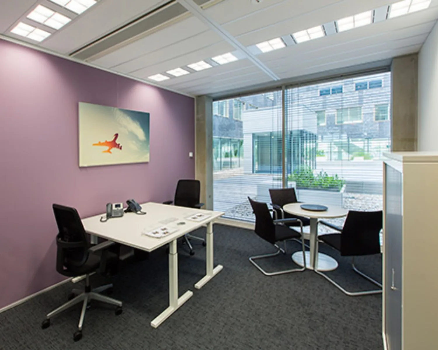 Modern ingericht kantoor met paarse muur, een bureau met stoelen, een ronde vergadertafel en een foto van een vliegtuig aan de muur.