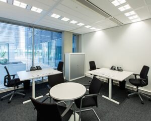 Moderne kantoorruimte met vier bureaus, vergadertafel en grote ramen met jaloezieën.