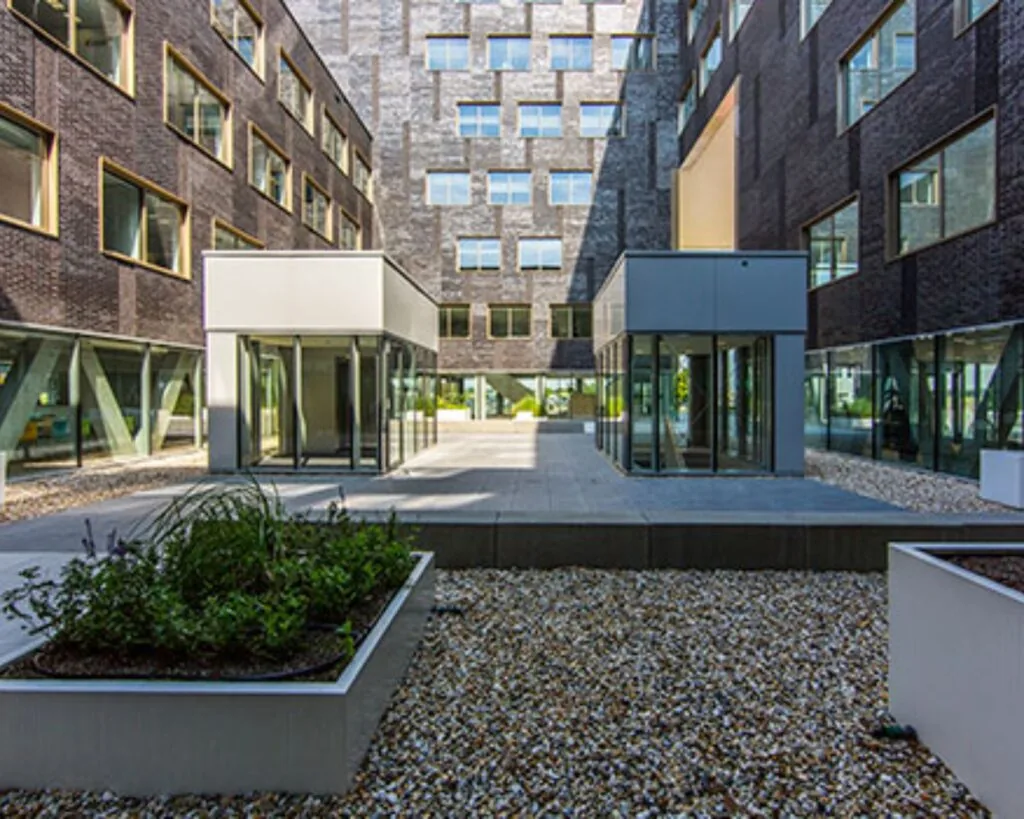 Binnenplaats van een modern kantoorpand met glazen entrees, grijze bestrating en verhoogde plantenbakken.
