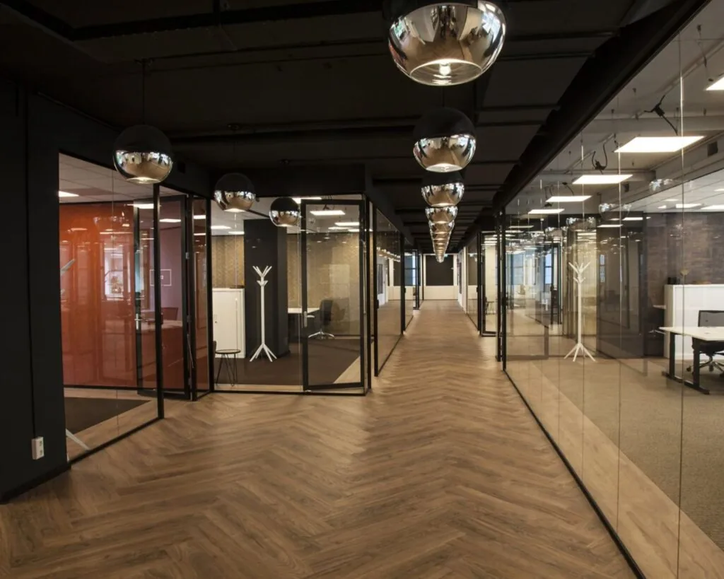 Moderne kantoorruimte met glazen wanden, houten visgraatvloer en spiegelende hanglampen.
