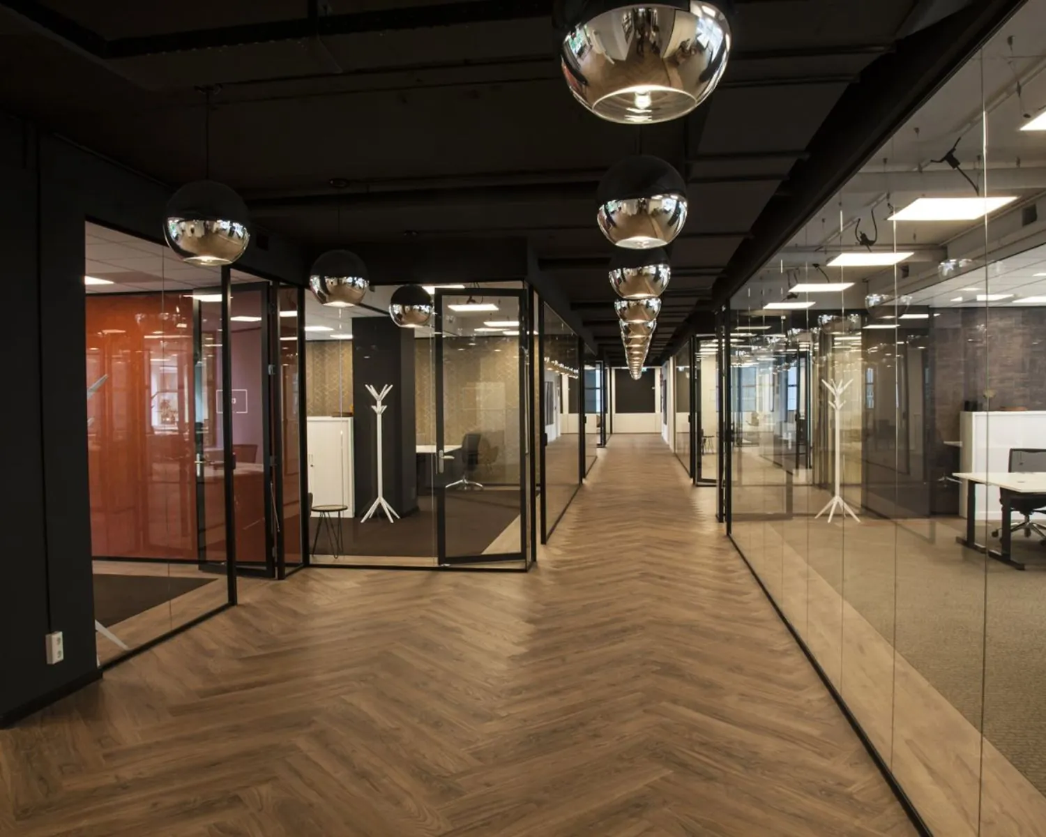 Moderne kantoorruimte met glazen wanden, houten visgraatvloer en spiegelende hanglampen.