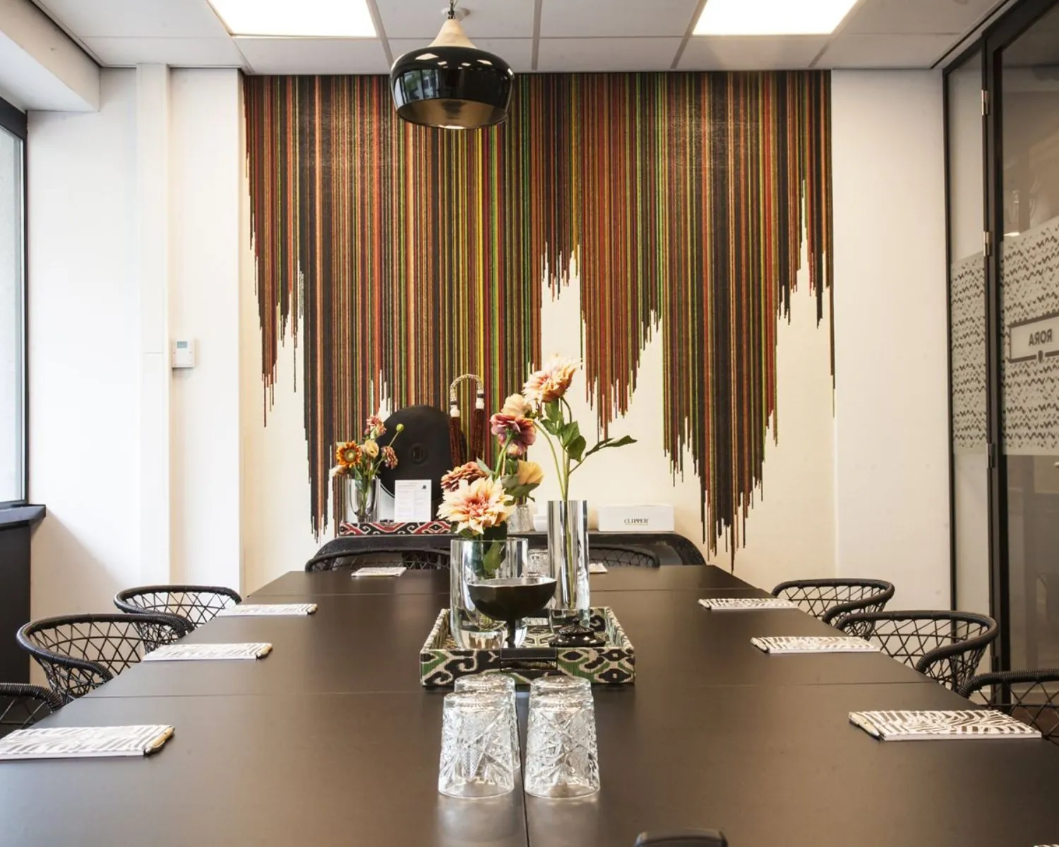 Vergaderruimte met een lange tafel, bloemen in vazen en een kleurrijke wanddecoratie van verticale strepen.