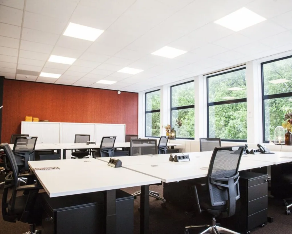 Moderne, open kantoorruimte met witte bureaus, zwarte bureaustoelen en grote ramen met uitzicht op groene bomen.