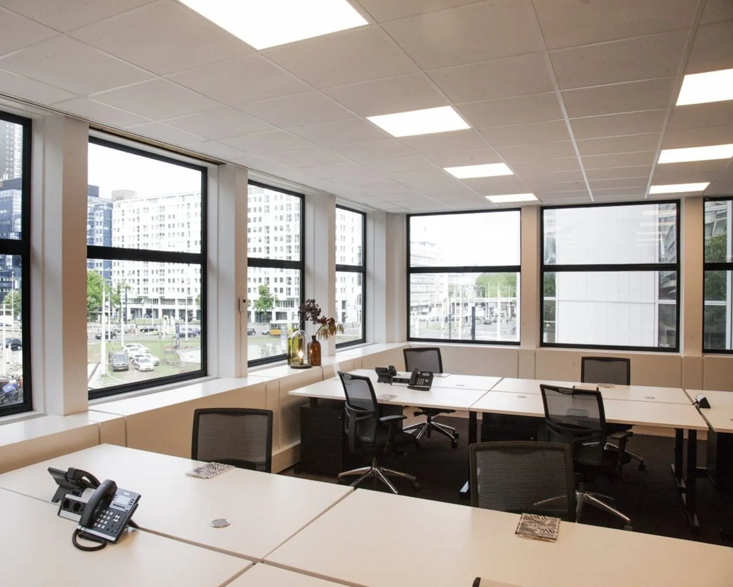 Moderne, lichte kantoorruimte met bureaus, bureaustoelen en uitzicht op een stedelijke omgeving met hoge gebouwen.