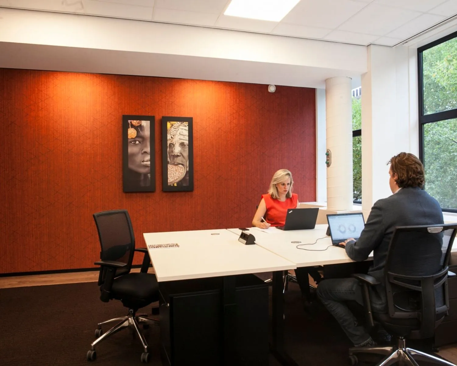 Twee mensen werken op laptops aan een vergadertafel in een modern kantoor met roodbruine wand en kunst aan de muur.