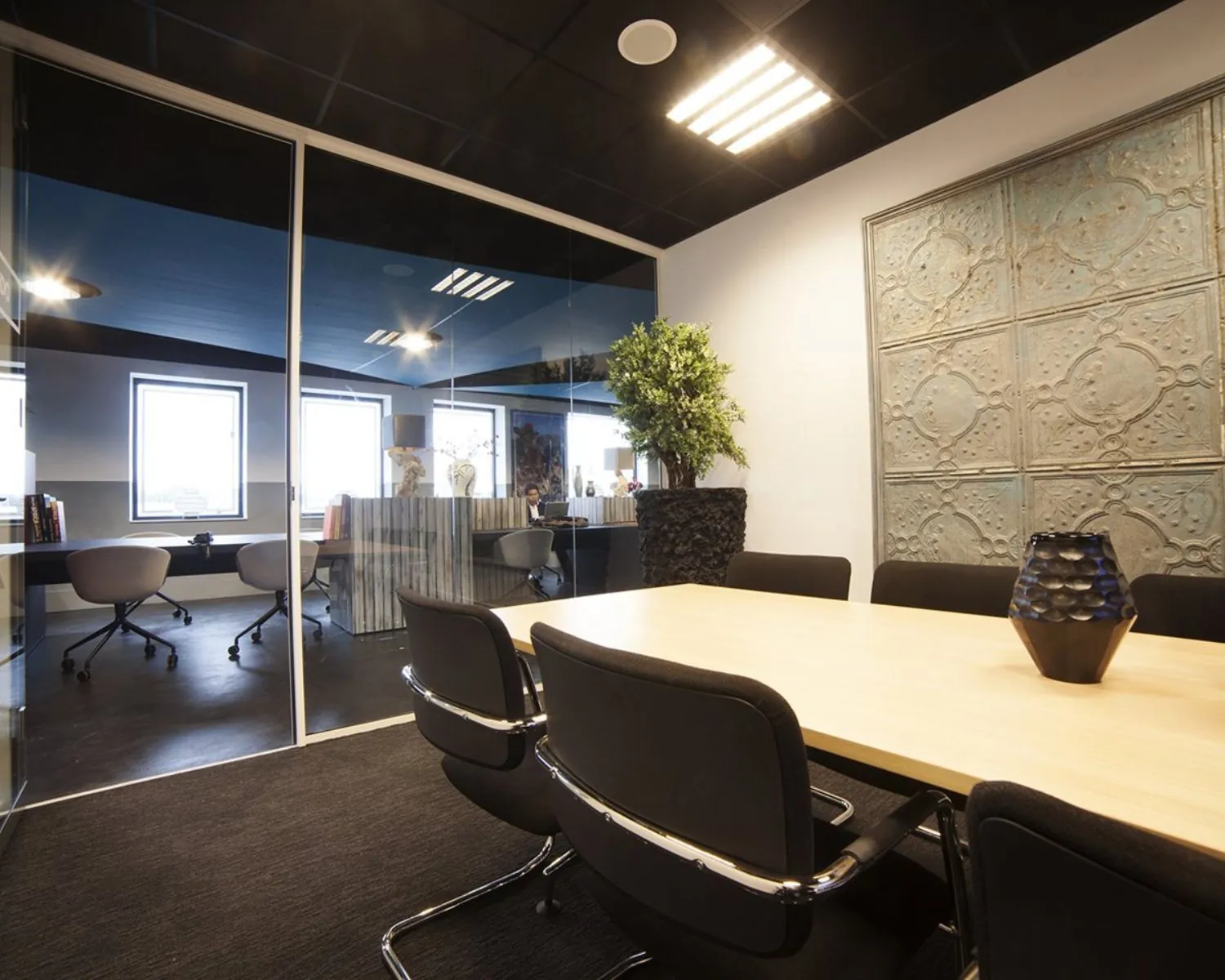Moderne vergaderruimte met zwarte stoelen, houten tafel, glazen wand en decoratieve elementen zoals een plant en een reliëfmuur.