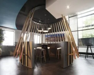 Moderne zithoek in een kantoorruimte met hoge barkrukken, hanglampen en houten lattenconstructie als afscheiding.